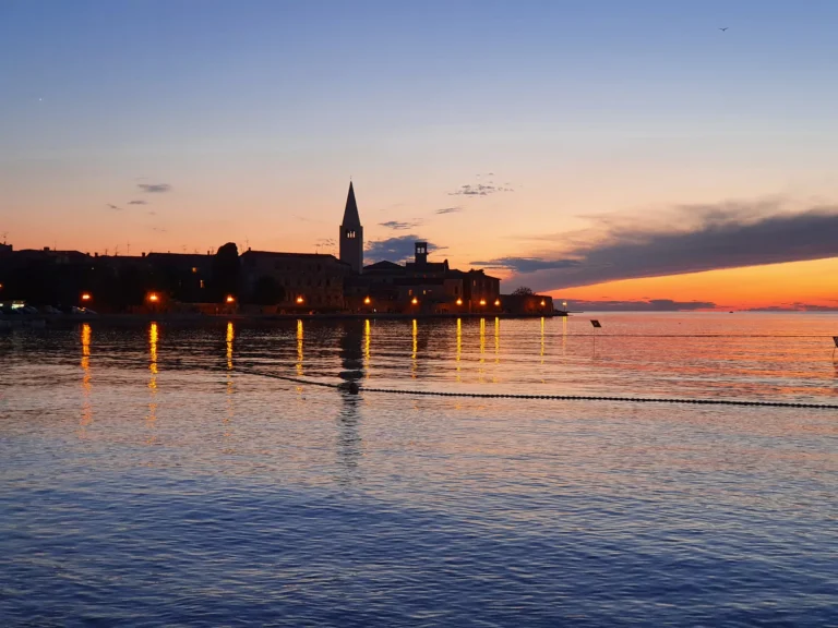A coastal town and church tower silhouetted against an orange sunset, with lights reflected on the water.