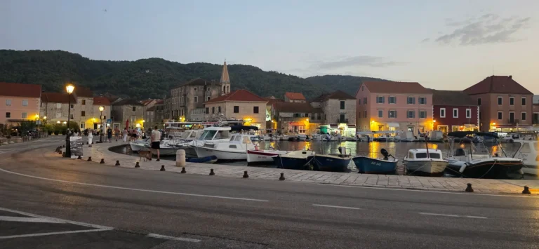 Boats moored beside a stone waterfront, with old buildings and wooded hills at dusk.