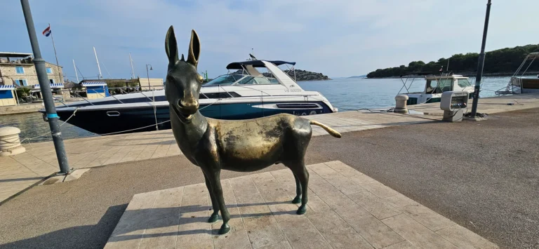 Bronze donkey statue on a sunny harbour promenade, with motorboats moored behind it