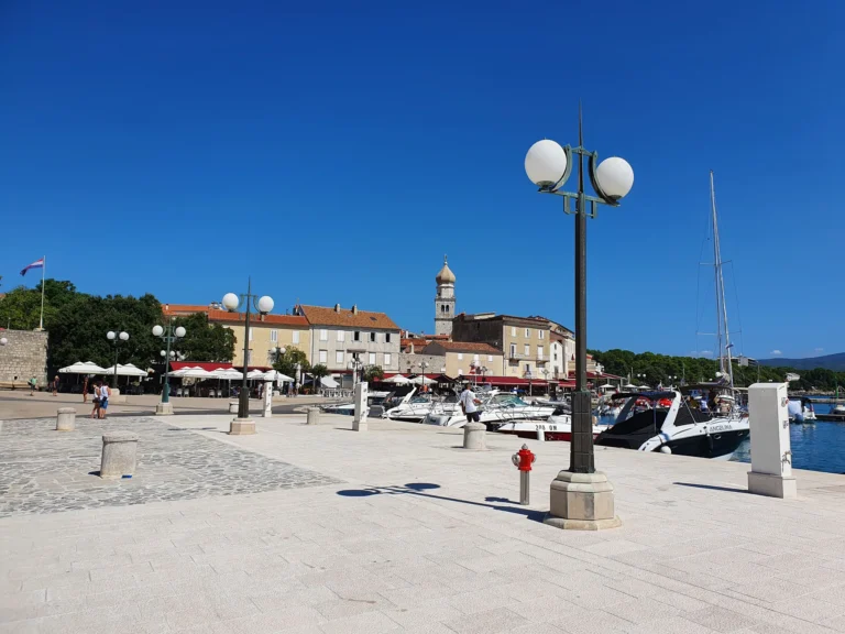 A sunny Croatian waterfront promenade with moored boats, stone buildings and a bell tower