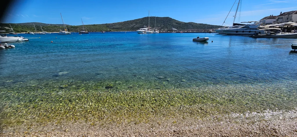 Clear blue water over a shallow pebble seabed, with boats moored beside a small coastal harbour.