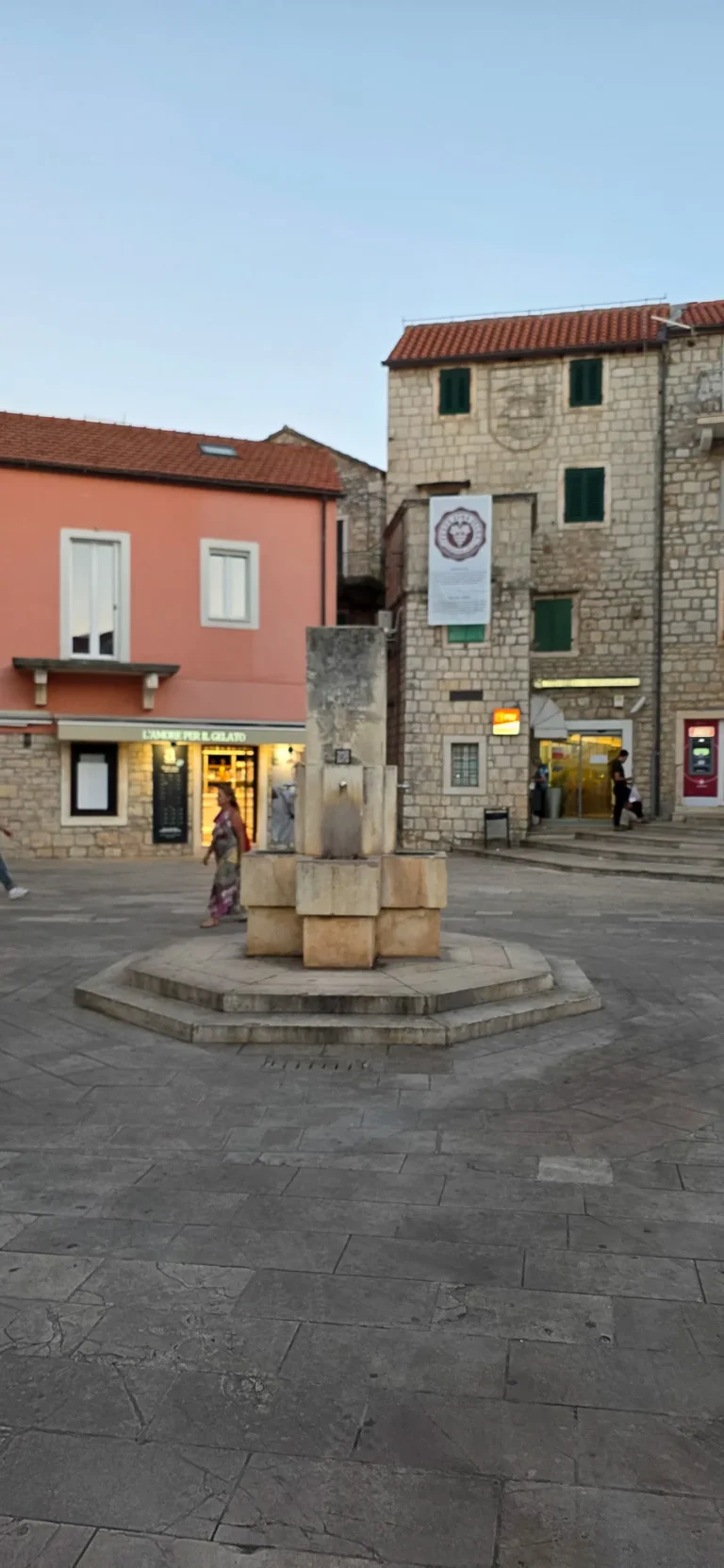 Stone fountain in a paved old-town square, with historic stone buildings and a pink storefront behind it