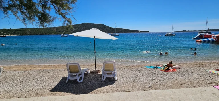 Pebble beach on the Croatian coast with swimmers, anchored sailboats and a white sun umbrella