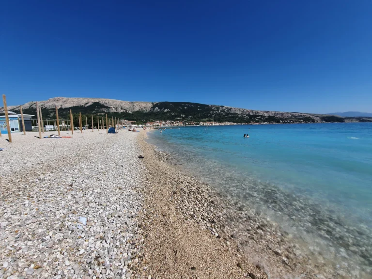 A broad pebble beach slopes into clear turquoise water beneath a rocky coastal ridge.