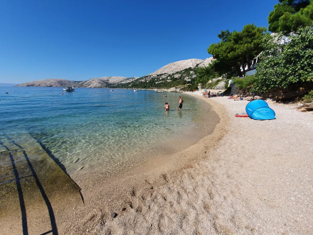 A pebbly Croatian beach curves beside clear shallow water, pine trees and rocky hills.