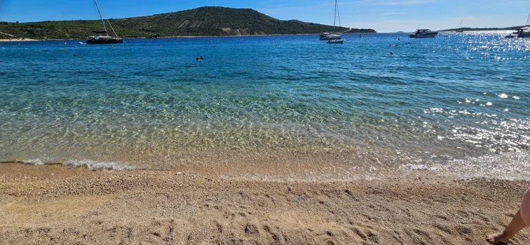 A pebbly beach meets clear blue water, with swimmers and boats offshore beneath a green coastal hill.