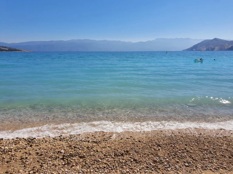 Pebble beach beside clear turquoise water, with distant mountains across the sea on the Croatian coast