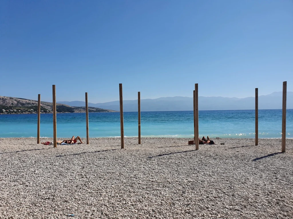 Sunbathers on a pebble beach beside clear blue water, with wooden posts and distant coastal mountains