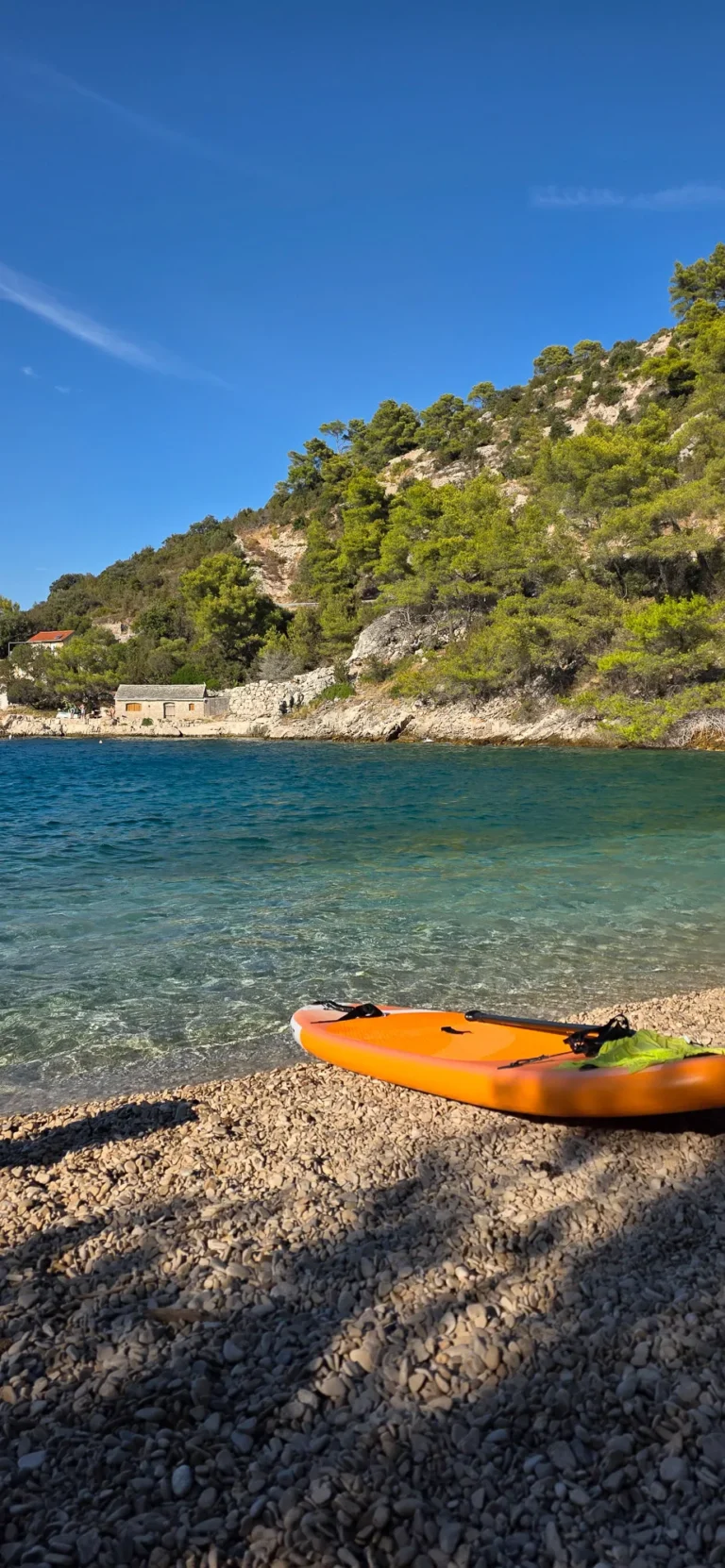 Orange kayak on a pebble shore beside clear water and a pine-covered rocky coast in Croatia