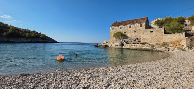 A clear pebble cove beside a weathered stone waterfront building, with a swimmer in the water.