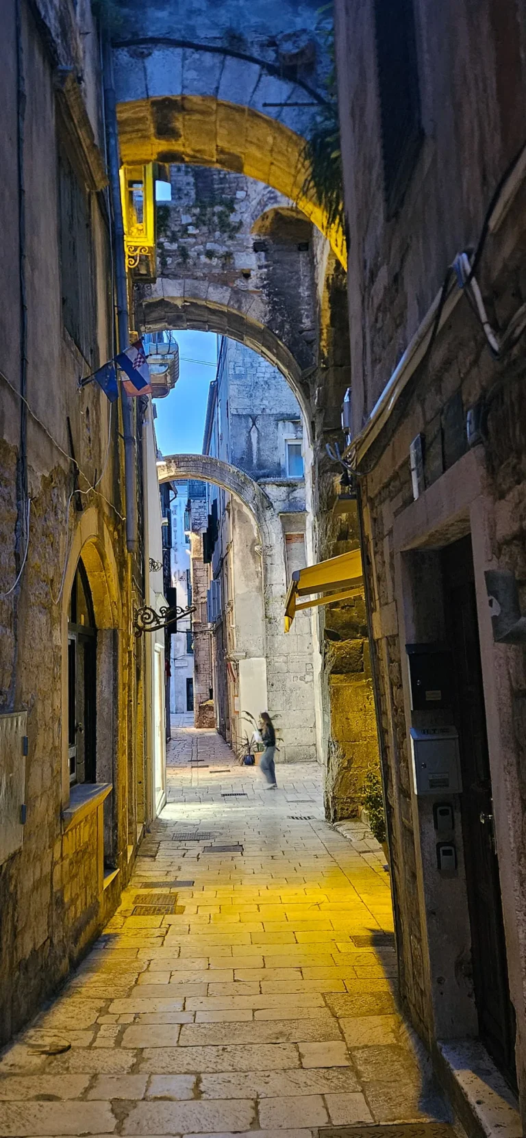 Narrow stone alley with overhead arches, warm streetlights and a Croatian flag