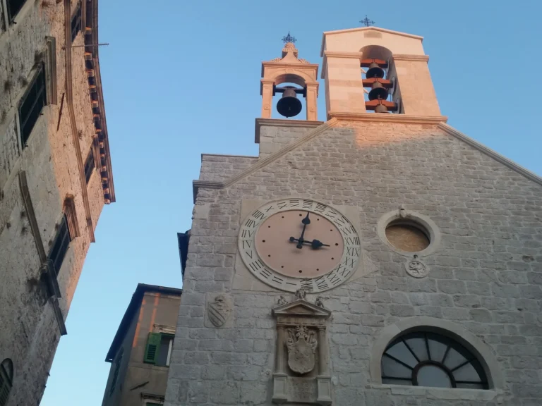 Stone church facade with a large Roman-numeral clock and two open bell towers against a clear blue sky