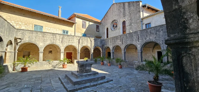 Stone courtyard with arched cloisters, potted palms and a central well beneath a bright blue sky