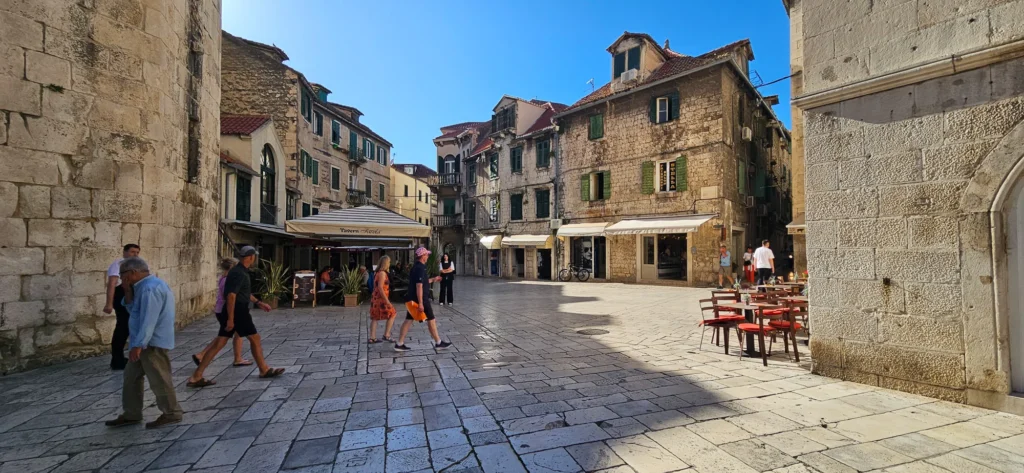 People cross a sunlit stone square lined with old-town buildings and outdoor café tables in Croatia.