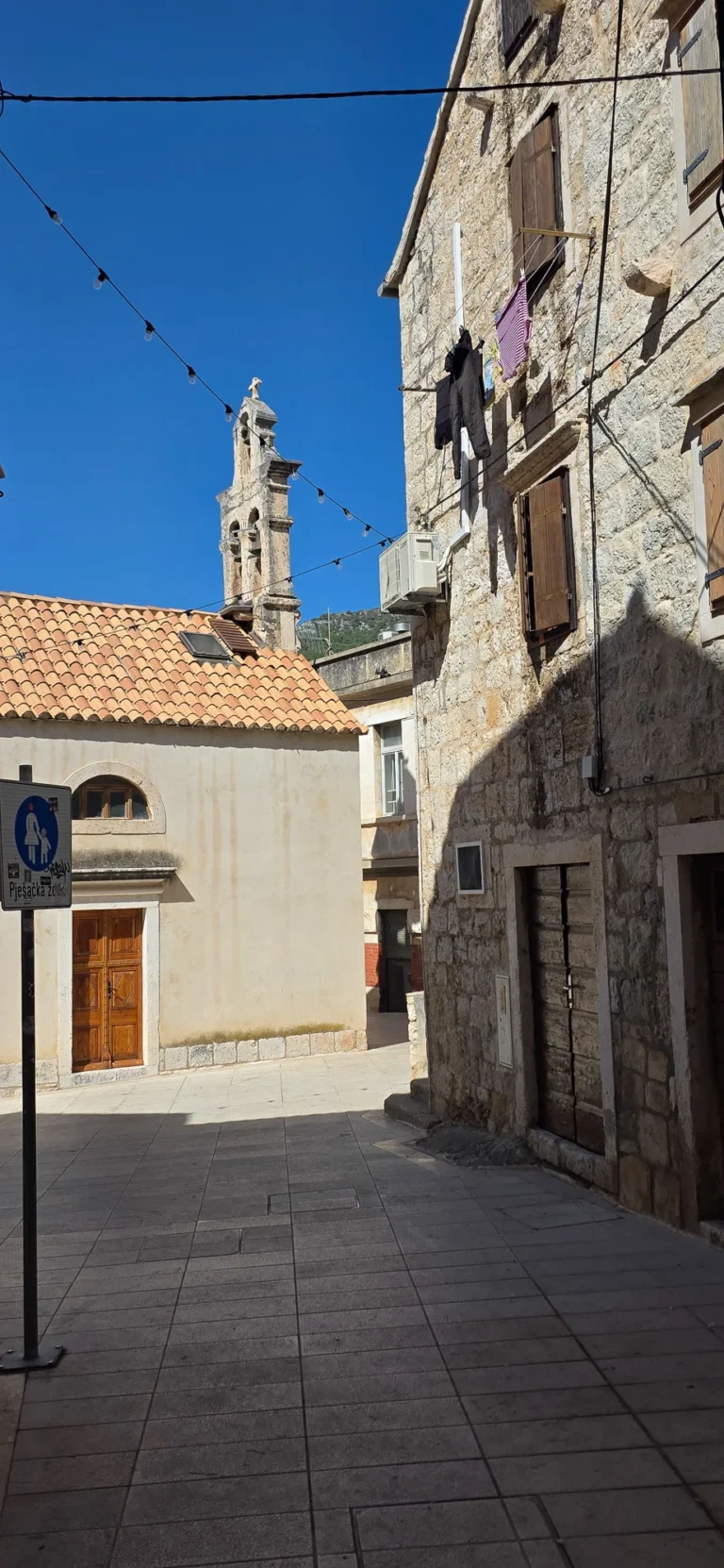 Stone buildings and a church bell tower frame a quiet paved street beneath a clear blue sky in Croatia.