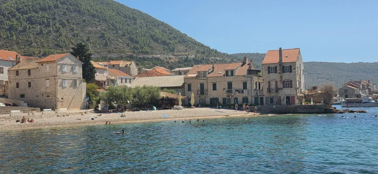 A small pebble beach below stone houses, with clear water and a wooded hillside behind
