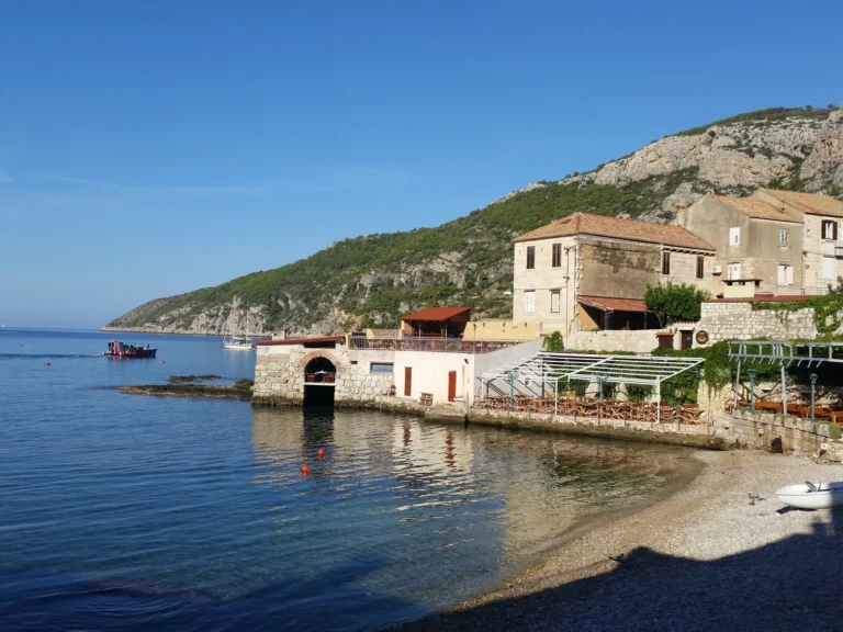 Stone waterfront buildings and small boats beside a calm pebble shore on the Croatian coast