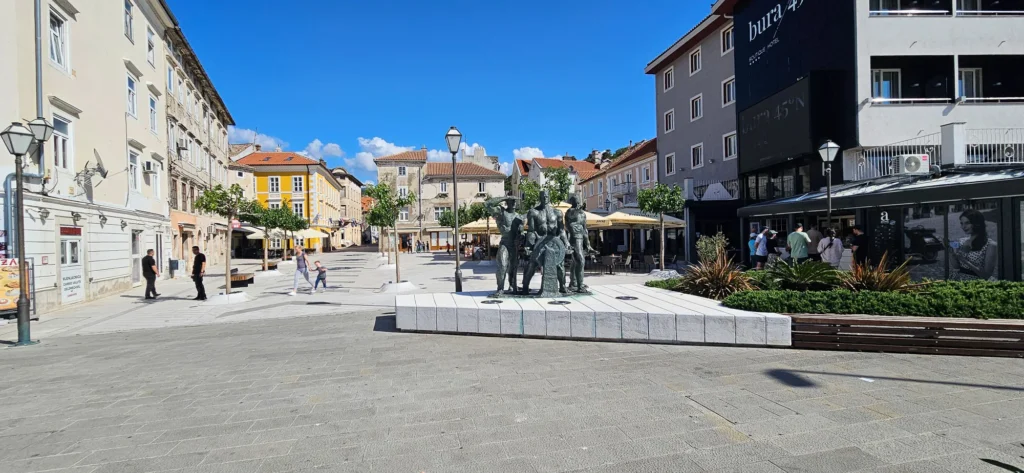 A paved Croatian town square with a bronze sculpture, historic buildings and outdoor café seating.