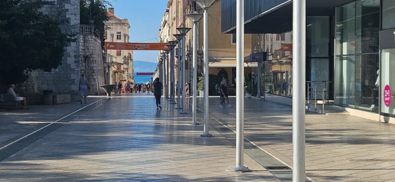 A broad tiled pedestrian promenade leads toward the sea, between old stone walls and modern glass-fronted buildings.