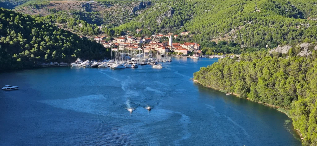 Boats in a blue-water harbour below a village and densely wooded hills in Croatia