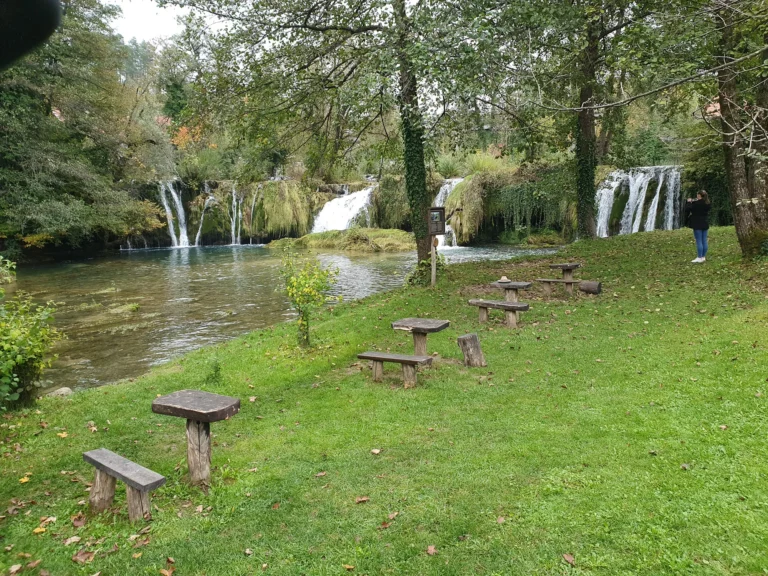 A series of waterfalls flows into a river beside a grassy woodland bank with rustic wooden stools.