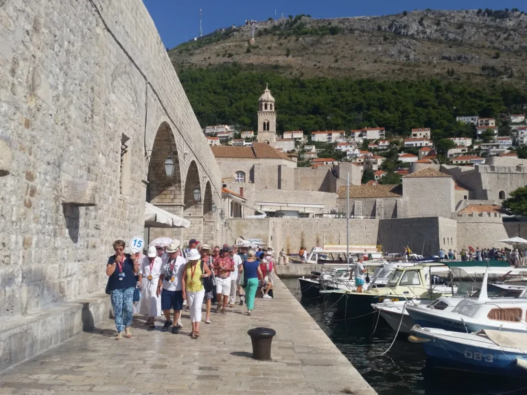 Boats line Dubrovnik Old Town harbour beside stone walls and a busy waterfront walkway.