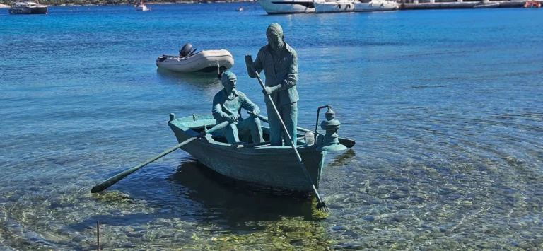 Two sculpted fishermen sit and stand in a small boat in clear, shallow coastal water.