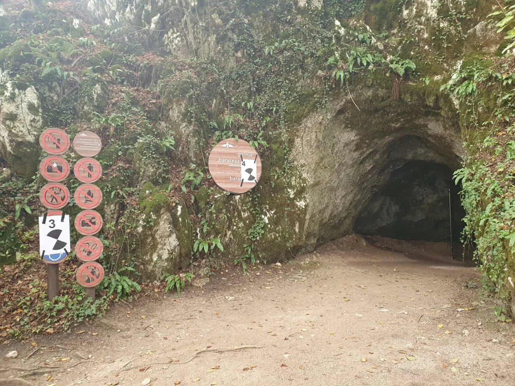 The leafy, rocky entrance to Gornja Baraćeva Cave, with visitor signs beside the path.