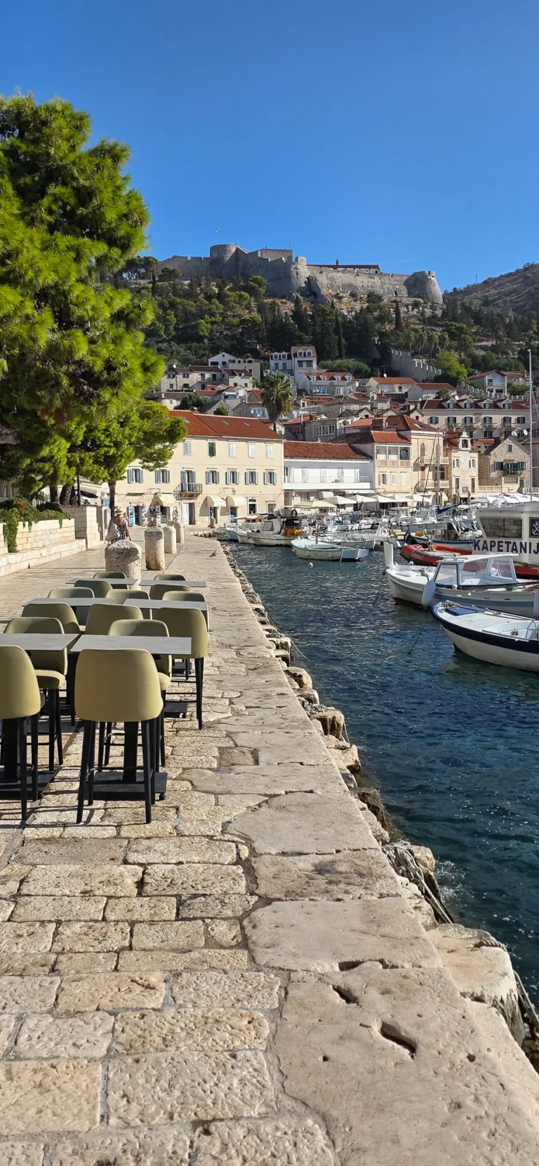 Boats line Hvar Town’s harbour below the hilltop fortress, with a stone waterfront promenade in the foreground.