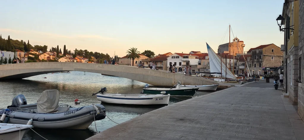 Boats moored beside the bridge and stone waterfront buildings in Jelsa harbour at sunset.