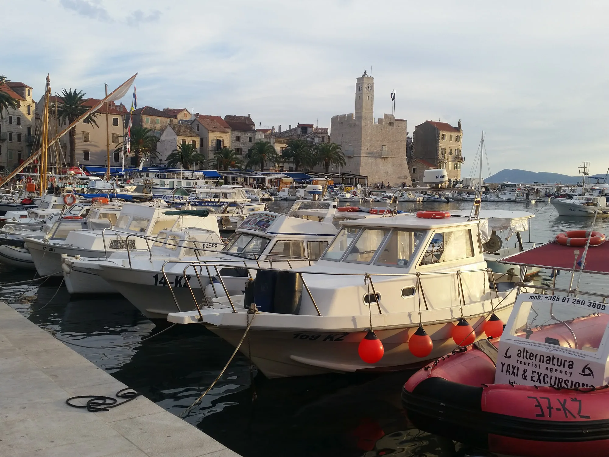 Excursion boats moored in Komiža harbour, with the stone clock tower and waterfront buildings behind them.