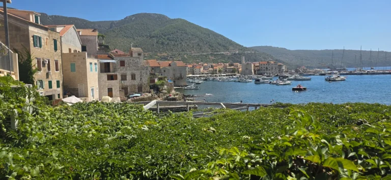 Stone waterfront buildings and boats line Komiža harbour, with a green mountain rising behind the town.