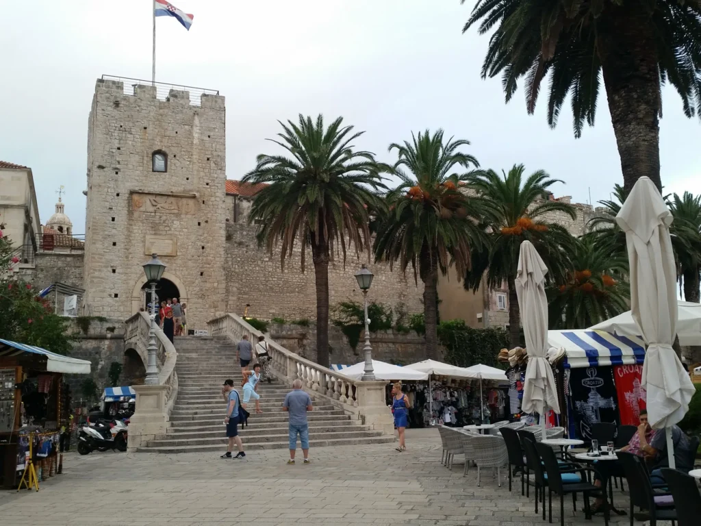 Stone steps lead to Korčula Land Gate, framed by palm trees and cafés in the old town.