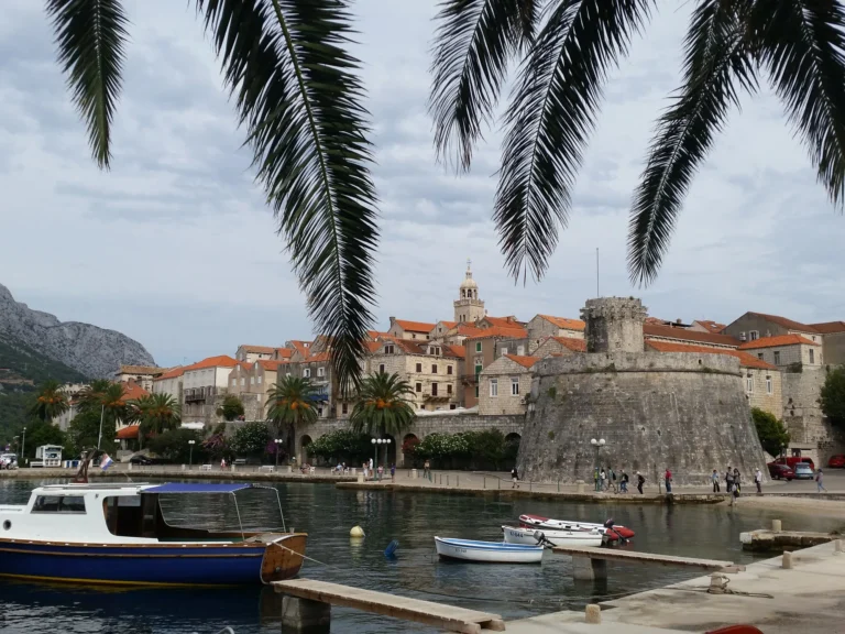 Korčula old town waterfront and stone tower, framed by palm fronds with boats moored in the harbour.