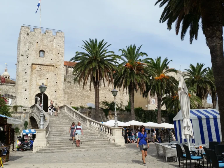 Stone steps lead to the arched entrance of Revelin Tower in Korčula Old Town, framed by palm trees.