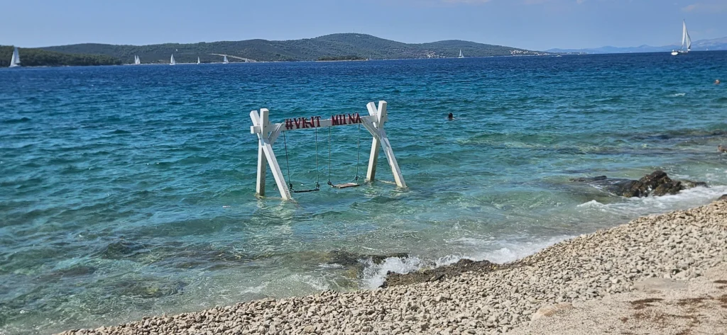 A wooden swing frame stands in the clear sea beside a pebble beach in Milna, Brač Island.