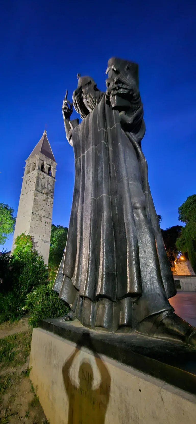 The bronze statue of Grgur Ninski beside a stone bell tower in Nin, viewed from below at night.