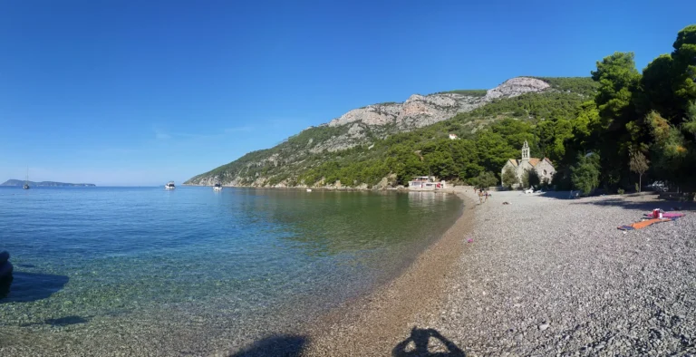Clear water and a pebble beach beneath wooded hills, with a small stone church by the shore.