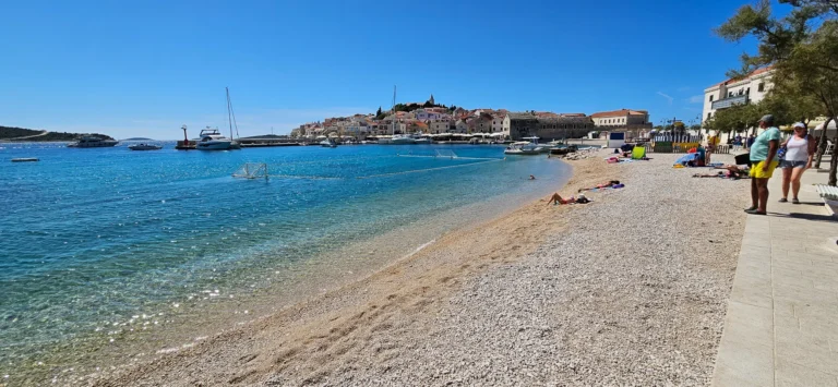A pebbly beach beside clear water, with boats in the harbour and a coastal town across the bay.