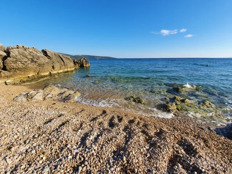 A pebble beach meets clear blue sea beside rocky outcrops on the Croatian coast.