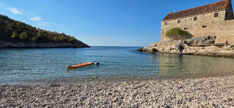 A pebble beach and clear cove with a stone building on the shore and a small kayak on the water