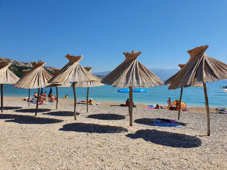 Thatched straw parasols shade beachgoers on a pebble beach beside clear blue water.