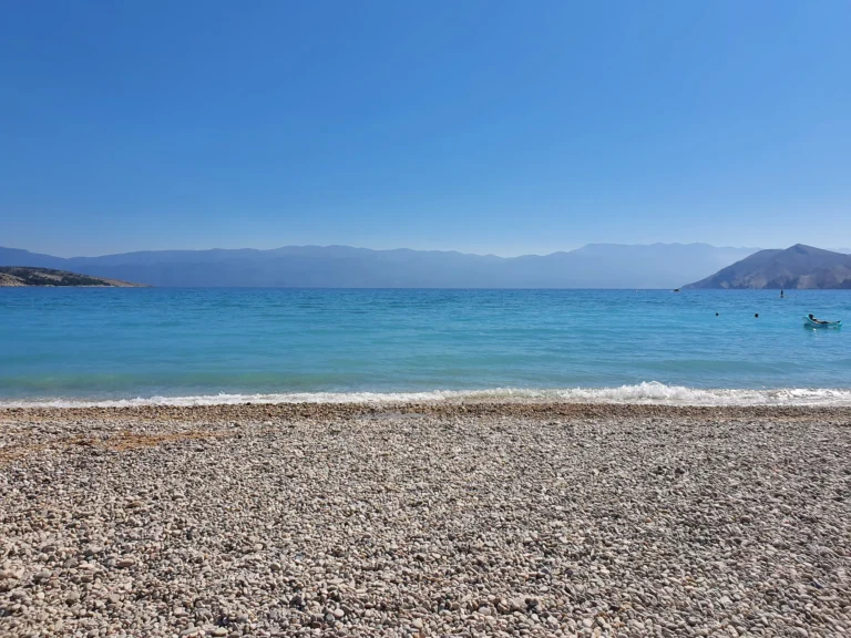 A broad pebble beach meets clear turquoise water, with hazy mountains across the bay.