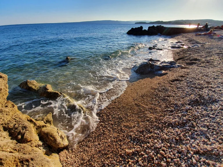 Small waves wash over a pebble beach beside rocks as people watch the sunset over the sea.