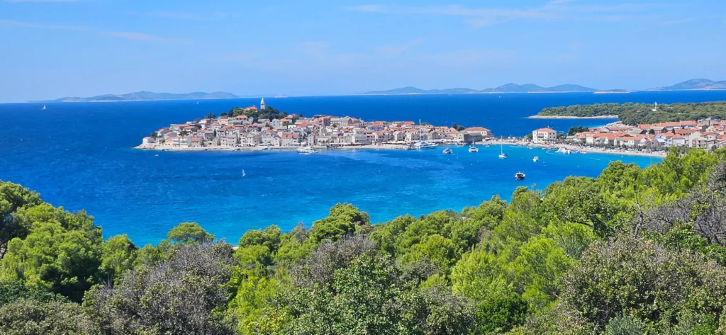 Primošten’s red-roofed old town sits beside a bright blue bay, framed by green coastal trees.