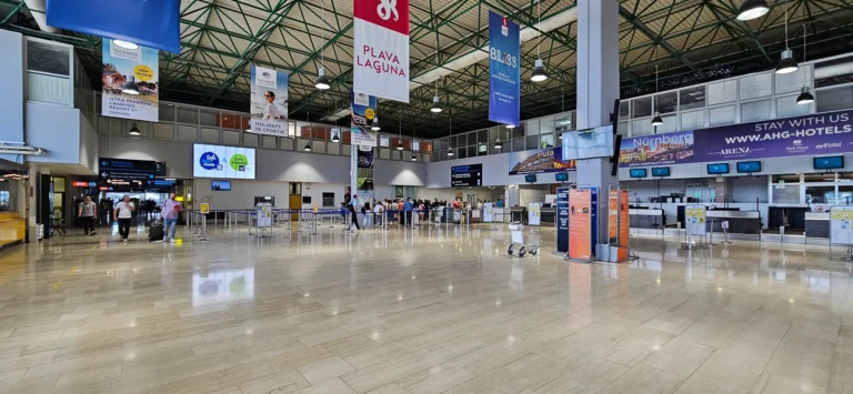 Check-in hall inside Pula Airport, with airline counters, overhead signs and passengers.