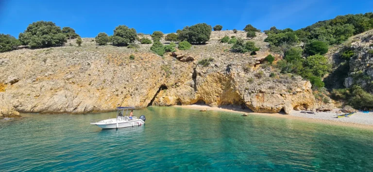 A small pebble cove beneath pale rocky cliffs, with clear turquoise water and a moored motorboat.