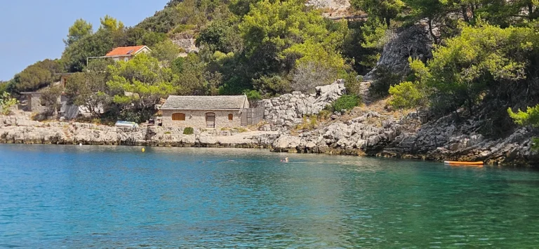 Clear blue water below a rocky cove with stone houses and pine-covered slopes.