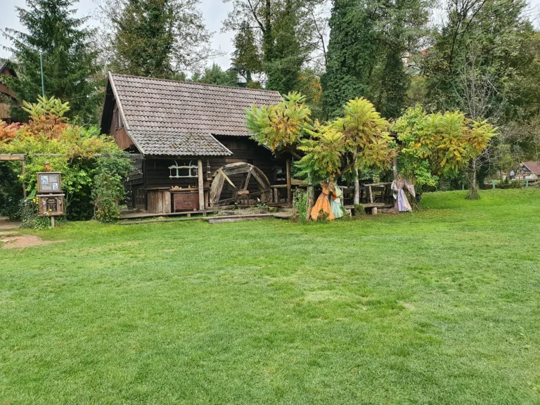 A rustic wooden building with a large water wheel, colorful garden figures and a broad green lawn.