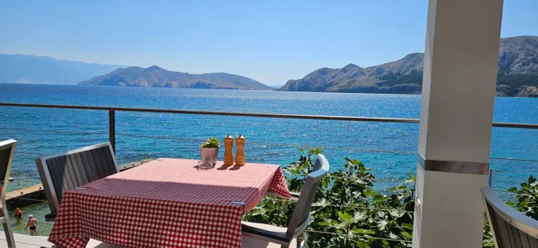 Red-check tablecloth and chairs on a terrace overlooking blue water and rugged coastal hills